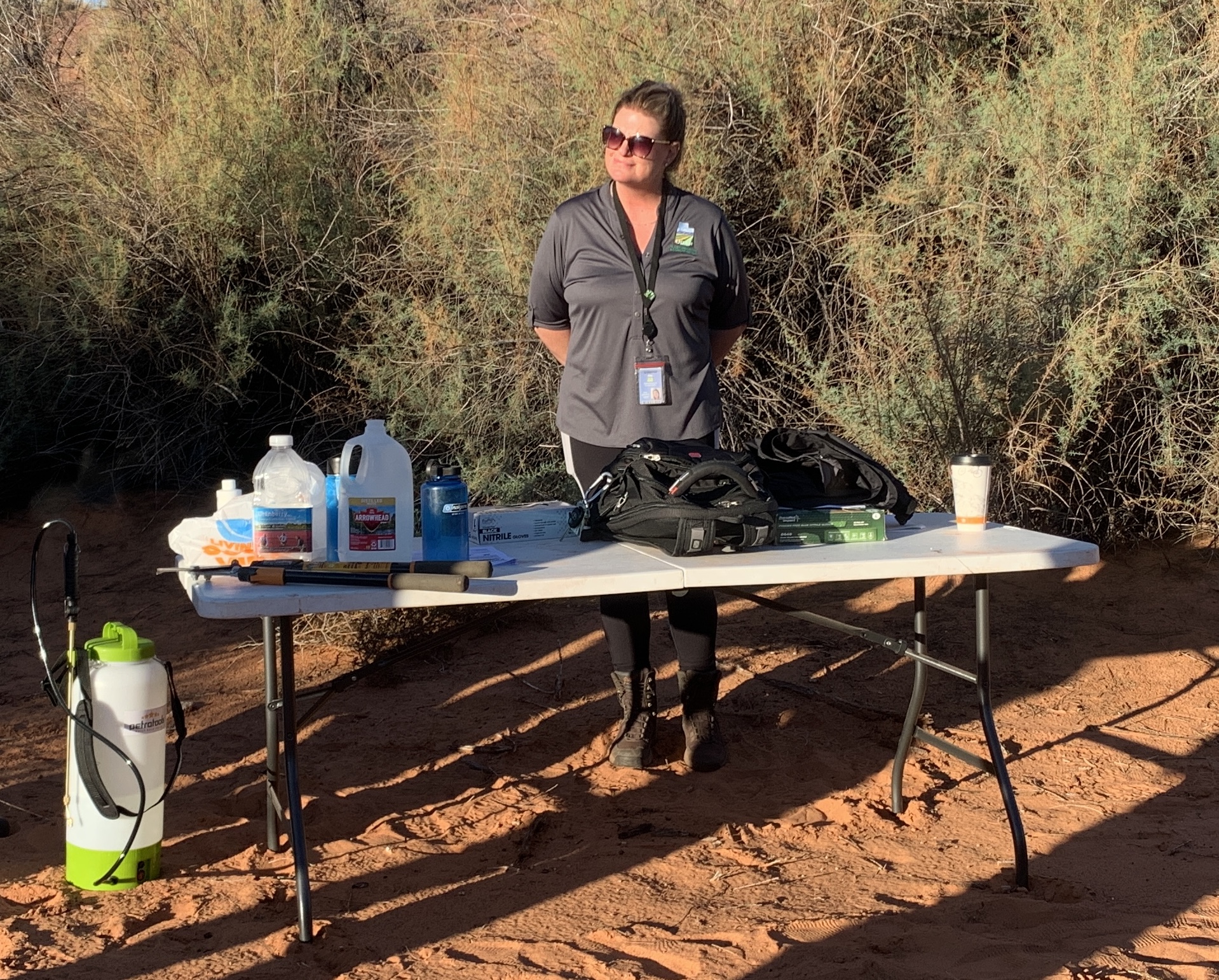 Amy davidson with Utah department of agriculture, stands at a table in a desert clearing in front of some tamarisk trees with some herbicide and other items spread out on the table