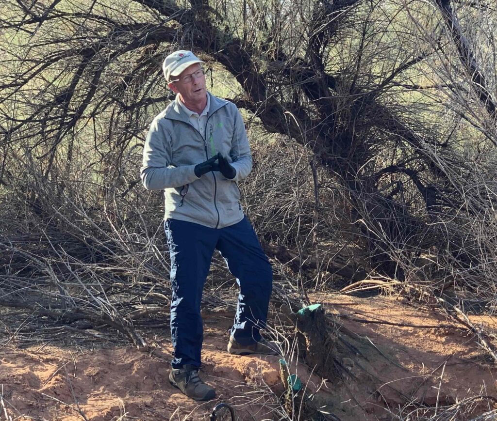 Richard Heflebower stands in front of a tamarisk in a clearing while talking to DPI volunteers about environmental conservancy work